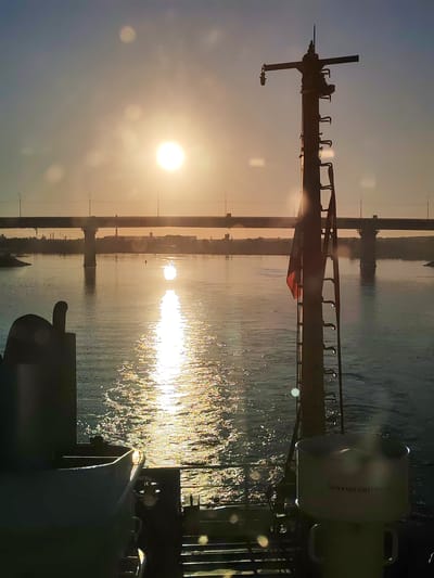 The image captures a stern-view perspective from a moving vessel on a wide river, likely in the city of Нифантово, Russia, during either sunrise or sunset. The sun, a bright, intense orb, is positioned in the upper right portion of the frame, casting a shimmering, golden reflection that extends vertically across the water towards the foreground.
In the midground, a long, multi-span road bridge, characterized by multiple concrete piers, stretches horizontally across the river, silhouetted against the bright sky. Small streetlights are visible atop the bridge. Distant shorelines with faint industrial or urban structures are discernible beneath the bridge, also in silhouette.
The foreground is dominated by elements of the vessel. On the right, a tall, dark mast or antenna structure rises, equipped with horizontal cross-arms near its top and a series of ladder-like rungs along its vertical axis. A small, red flag, partially illuminated, is visible on the mast. Below this mast, a light-colored, cylindrical object with handles is present, bearing the Cyrillic inscription "АККУМУЛЯТОРНАЯ" (Akkumulyatornaya), indicating "Battery Room" or "Battery Operated." On the far left, a dark, curved section of the vessel's superstructure or a vent is partially visible. The river's surface in the foreground shows discernible ripples and a wake, consistent with a vessel in motion. Multiple instances of lens flare are visible throughout the image due to the direct light source.