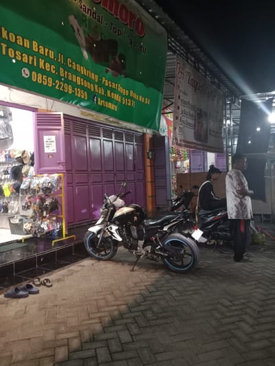 This image captures a nighttime street scene in Cangkring, Kendal Regency, Indonesia, illuminated by artificial light sources. The foreground consists of interlocking grey concrete pavers laid in a herringbone pattern.
On the left, a storefront with purple accordion-style security doors is partially open, revealing merchandise. A large green banner above the entrance advertises "Sandal - Topi - Tas - dll" (Sandals - Hats - Bags - etc.) and provides an address: "Jl. Cangkring - Pasar Srogo Ruko No. 04 Brangsang Kab. Kendal 51371". A phone number, "0859-2299-1359", and a Facebook handle, "f Artomoro", are also visible on the banner. Inside the open section of the store, various shoes, sandals, and hats are displayed on white shelving and a yellow metal rack. A black basket is positioned on the floor near the rack. Directly in front of the storefront, on the paved ground, are two blue slide sandals and two black flip-flops.
Two motorcycles are parked in front of the store, facing left. The motorcycle closest to the viewer is black and white, featuring blue accents on the rear wheel rim. Behind it, a darker motorcycle has a male seated on it. This individual wears a dark cap and a black long-sleeved shirt with a white stripe on the back, and is looking towards the right. A third motorcycle is partially visible behind the second one.
To the right of the parked motorcycles, a second male stands, facing right. He wears a light-colored, possibly patterned, t-shirt and dark pants with red stripes on the sides. His right arm is bent at the elbow. Further to the right, a third male is partially visible, wearing a dark shirt.
Above the standing men and motorcycles, a second, partially obscured banner is visible. It advertises "Nyewakan Kostume Profesi Baju Adat Anak Dewasa Dll" (Rents Professional Costumes, Traditional Children's and Adult Clothing, etc.). On the far right of the scene, a green gas cylinder is present on the ground. The sky is dark, indicating nighttime conditions.