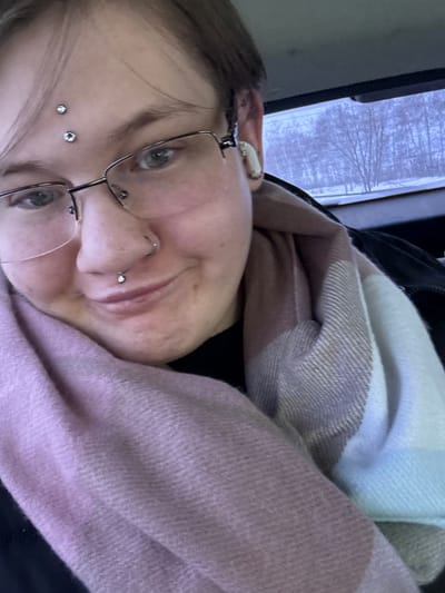A headshot-style image depicts an individual in what appears to be an indoor setting, possibly a vehicle, within the city of Ershovka, Russia. The individual has short, light brown hair, is wearing rectangular metal-framed glasses, and has blue-grey eyes. Visible facial piercings include two dermal piercings with silver studs on the forehead, a silver ring in the septum, and a silver stud in the philtrum. A white earbud is positioned in the right ear. The individual displays a subtle smile. Clothing consists of a black garment, partially visible at the neck, covered by a large, woven scarf featuring sections of light pink, light brown/beige, and light blue/white. Through a window in the background, a winter landscape with bare, snow-covered trees under an overcast sky is visible.