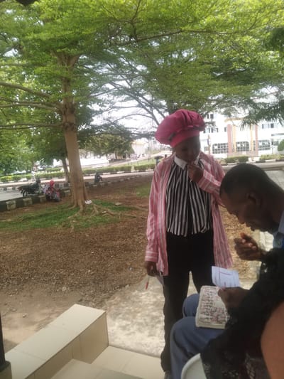 The image shows a woman and child standing outside in what appears to be a public space in Osu, Nigeria. The child is wearing a pink head covering, a striped shirt, and black pants, with a pink and white striped jacket or shirt over the striped shirt. An adult is standing nearby. There are papers in the adult's hand. They are standing on a surface that appears to be concrete or stone, near a curb. A tree with green foliage is in the background, as is a multistory building. The overall environment is well-lit, suggesting it is daytime.