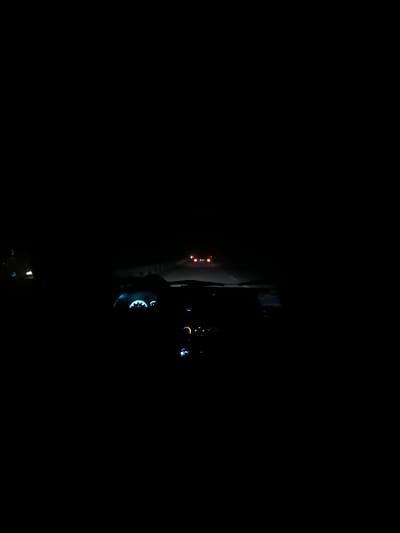 A nighttime view from inside a vehicle. The dashboard gauges emit a faint blue glow. Further ahead, the headlights of another car shine faintly on the road. The overall scene is very dark, making it difficult to discern details beyond the immediate vicinity. The dark road stretches into the distance, guided by the weak illumination.