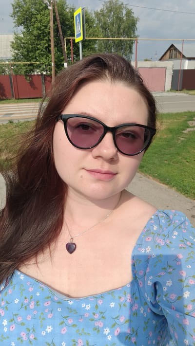 A medium shot captures a young woman outdoors on a sunny day. She is wearing dark-rimmed sunglasses and a light blue dress with a floral pattern of pink and white flowers. A silver necklace with a heart-shaped pendant rests on her chest. Her long, brown hair is slightly windswept.
The background shows a street with a pedestrian crossing, a road, and green grass. Trees and buildings are visible further in the distance. A yellow and blue pedestrian crossing sign is present on a wooden pole. The sky appears cloudy. The overall impression is that of a casual, everyday moment, likely in a suburban or rural setting in Krupskoy, Russia. No text is visible in the image.