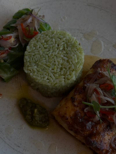 A close-up, overhead shot displays a plated meal on a textured, light-colored ceramic dish in Belalang, Indonesia. The centerpiece is a golden-brown piece of seared fish or poultry, topped with a relish composed of thinly sliced shallots, red chili, and fresh green microgreens. To the left of the main protein is a cylindrical mound of light green-colored rice. Further to the upper left, a portion of green leafy salad, intermixed with sliced shallots and red chili, is visible. A small dollop of dark green, textured sauce or paste is present on the lower left of the plate. The plate exhibits several oil or sauce streaks and speckling. No individuals are present in the frame.