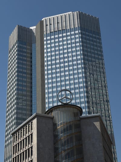 This is a low-angle, Dutch-tilted shot of a modern skyscraper, likely in Frankfurt, Germany, against a clear blue sky. The skyscraper is clad in glass and metal panels, with a distinctive pattern of alternating light and dark squares creating a checkerboard effect on some sections. The building's architecture is characterized by sharp, geometric lines and a sleek, reflective facade.
A prominent detail is the Mercedes-Benz logo, a three-pointed star within a circle, visible on a cylindrical element attached to the side of the building. This suggests the building is either headquarters or a significant office space for Mercedes-Benz or a related entity. The building's orientation, viewed from below and tilted, emphasizes its height and grandeur.
The lighting suggests it is daytime, possibly late morning or early afternoon, given the strong shadows cast by the architectural elements. The clear sky indicates fair weather. The scene is devoid of people, focusing solely on the architectural subject and its prominent branding. The overall impression is one of modern corporate architecture and a well-established automotive brand. No visible text other than the brand logo is present in the image.