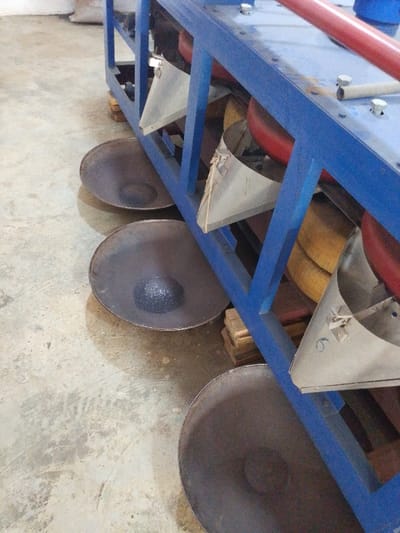 The image displays an industrial processing scene located in Olomoda, Nigeria. A prominent blue metal framework, consistent with heavy machinery, occupies the right and central portions of the frame. This structure is positioned on a light-colored concrete floor, with stacked wooden blocks visible beneath it, possibly for support or leveling.
Below the blue framework, three dark, shallow, circular metal pans are arranged in a diagonal line on the floor. The middle pan contains a small, distinct conical pile of dark, granular or crystalline material. The other two visible pans are empty.
Attached to the blue framework are multiple inverted, silver-colored conical or pyramidal funnels, designed to direct material downward. One of these funnels features a faint numeral "6" inscribed near its base. Behind these funnels and integrated within the blue framework, several large, red, cylindrical objects are partially visible, alongside coiled sections of yellow or brown material, possibly hoses or belts.
The concrete floor exhibits scuff marks and minor discoloration, indicating an active industrial environment. In the blurry background, an indistinct white sack is partially visible. No individuals are present in the depicted scene.
