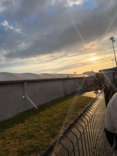 The image depicts a cloudy sunset scene in Tillé, France. The sun, low in the sky, casts a warm glow, creating sunbeams that pierce through the clouds. In the foreground, a dark metal fence runs diagonally across the frame, its wires casting long shadows on the asphalt path behind it. To the left of the fence, a grassy area with dappled sunlight stretches towards a long, white, tent-like structure with a corrugated roof. A white downspout extends from the structure to the ground. Further down the path, more tents and some figures are visible, along with a tall pole with lights or speakers. The overall impression is of an outdoor event or venue bathed in the golden light of a setting sun.