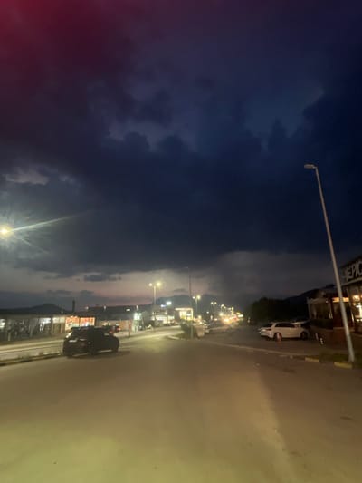 The media file depicts a street scene at dusk or dawn in Dub, Montenegro. The sky is a deep blue, with dramatic clouds and streaks of pink and purple from the setting or rising sun. Streetlights are beginning to illuminate the road, which appears wet, suggesting recent rain or mist.

The scene is a street lined with buildings on the left side, with various shops and businesses visible, some illuminated by interior lights. A white car is parked on the side of the road. Further down the street, more lights indicate continued development or activity. On the right side of the road, there's a dark-colored vehicle with a person on it, potentially a motorcycle, parked or moving slowly. The road itself is paved and appears to be a main thoroughfare.

The overall mood is somewhat atmospheric and tranquil, characteristic of the transition between day and night. The wetness of the road and the dramatic sky contribute to a slightly moody yet serene feeling. There are no prominent emotional expressions visible on any people, as they are either distant or obscured. No discernible text is visible in the media file. The time of day is clearly indicated by the lighting conditions and the sky's colors.