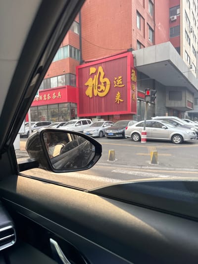 The image captures an urban commercial street scene in Huanggu District, China, viewed from the interior of a vehicle during late afternoon or early evening, characterized by warm, golden sunlight. The foreground shows the black interior door panel and window frame of the vehicle, with the side mirror reflecting a person, presumably the driver, wearing a white face mask.

Beyond the vehicle, multiple sedans of varying colors, including silver, black, and white, are parked along the street. The street features yellow-tipped concrete bollards and white crosswalk markings. A red traffic light, illuminated, is visible on the right side.

In the background, a multi-story building with a reddish-brown exterior facade and numerous rectangular windows dominates the scene. A large, prominent vertical red sign with golden Chinese characters "福运来" (Fúyùnlái), meaning "Fortune/Good Luck Comes," is affixed to this building. Below it, a smaller horizontal red sign displays white Chinese characters "白酒茶具" (Báijiǔ Chájù), indicating a store selling liquor and tea sets. A gray, flat cantilevered overhang extends from the building structure on the right side.