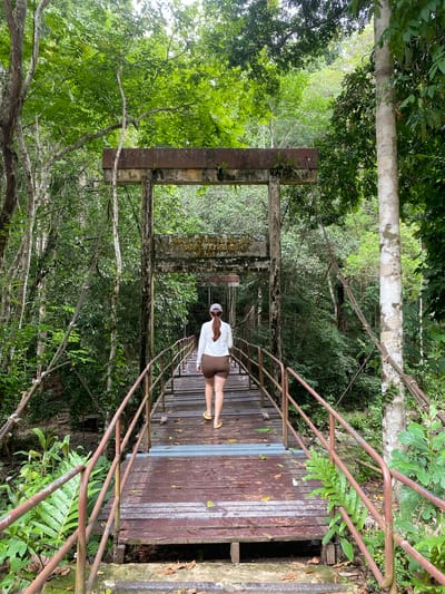 A medium shot captures a woman standing on a wooden suspension bridge within the lush greenery of Tambon Thai Mueang, Thailand. The bridge is constructed with brown wooden planks, bordered by rusty metal railings. The woman, positioned slightly off-center, is dressed in a light-colored jacket and shorts, complemented by yellow sandals. Above the bridge's entrance, a weathered sign displays text in Thai. The surrounding environment is densely vegetated with trees and foliage, casting diffused light throughout the scene. The camera angle offers a perspective looking down the bridge, enhancing the sense of depth within the image.