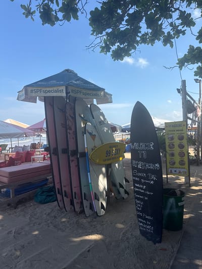 The image depicts a beach scene in Kuta, Indonesia, likely in the morning or afternoon judging by the lighting. In the foreground, a collection of surfboards are leaning against a wooden structure. A black surfboard with white lettering serves as a menu board, listing "WELCOME TO ELY & KIBANG SUNSET BAR," followed by drink and food items like "ICE COLD BINTANG," "RADLER," "SMIRNOFF ICE," "SPRITE," "COKE," "FOOD," "CHIPS & SNACK," "COCONUT ICE," "SOFT DRINKS," and "SUN BED." Adjacent to this, a yellow oval sign reads "WELCOME TO 99 SEXY SURF BOARD RENTAL & LESSON." Behind the surfboards, umbrellas provide shade for beach loungers and tables, with beanbag chairs visible. Further in the background, the ocean with small waves and a few distant surfers can be seen. A food menu sign for "99 WARUNG" is also visible. The environment is sandy, with some foliage from trees overhead casting shadows. There are no visible people actively engaged in activities, though beachgoers are suggested by the presence of loungers and umbrellas.