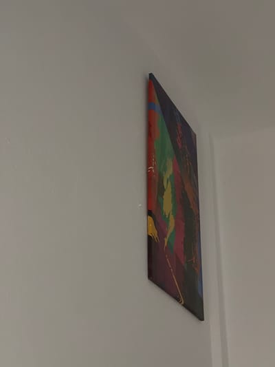 This is a medium shot, angled photograph of a piece of abstract art mounted on a wall. The artwork is rectangular and appears to be a canvas print or painting. It features bold, geometric shapes and abstract brushstrokes in a variety of colors, including shades of purple, red, green, yellow, and dark blue. The composition is dynamic, with diagonal lines and intersecting planes creating a sense of movement. The colors are vivid and contrast with the neutral gray of the wall. The artwork is positioned slightly off-center within the frame, with a significant portion of the wall visible above and below it. The lighting is even, suggesting an indoor setting. There is no text visible in the image. The scene depicts a decorative element within a room.