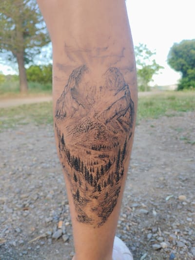 The image depicts a person's lower leg with a tattoo of a mountain landscape. The tattoo is black and white and features two prominent mountain peaks, a dense forest of pine trees, and a winding path at the bottom. The background of the image shows a gravel path and trees, suggesting an outdoor location. Based on the location in the city of Rubí, Spain, it's likely the photo was taken in a park or natural area within the city limits.