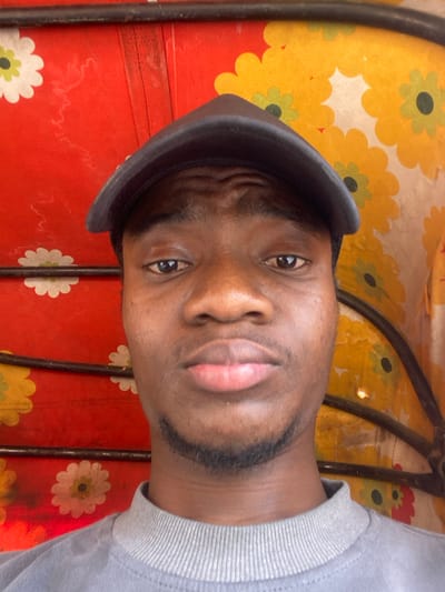A young adult male, appearing of African descent, is depicted in a close-up, head-and-shoulders portrait shot, likely within the interior of a vehicle in Baganakwo, Nigeria. He wears a dark grey baseball cap and a light grey crew-neck sweatshirt with a ribbed collar. His gaze is directed forward towards the camera, maintaining a neutral expression. His facial features include dark skin, dark eyes, full lips, and short dark stubble along his jawline and chin. The background consists of a brightly patterned fabric, predominantly red on the left and yellow on the right. Both sections feature a repeated floral motif: white flowers with light green centers on the red background, and light green-centered flowers on the yellow background. Two horizontal dark metal bars traverse the background, indicative of structural components within the vehicle's canopy or frame. A vertical seam is visible on the red fabric to the left of the subject's head.