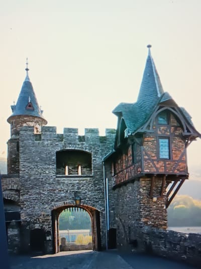 The image shows a view of a castle-like structure with a stone archway leading through it. To the left, there is a circular tower with a conical roof topped with a spire. To the right, a half-timbered building with a steeply pitched roof juts out from the stone wall. The structure appears to be made of stone and wood, with some areas featuring exposed brick. The archway is framed by wooden beams and has a gate with vertical slats. Beyond the archway, sunlight illuminates a treed area. The sky is overcast and bright. The scene does not depict the city of Serhiivka, Ukraine, but rather a medieval-style castle.