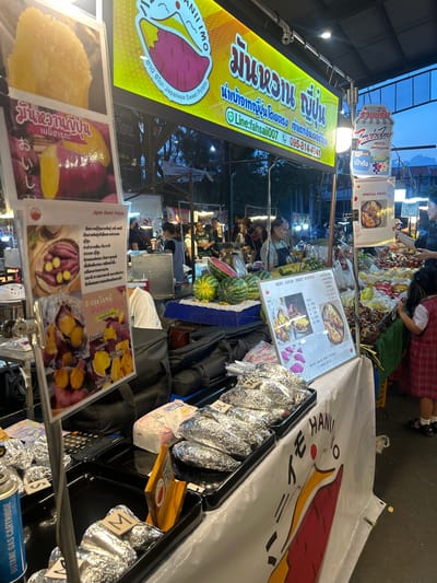 The image depicts an outdoor market stall in Tha Khlong Town Municipality, Thailand, specializing in Japanese sweet potatoes. A large, brightly lit yellow banner prominently displays text and an illustration of a sweet potato, indicating the name of the stall and contact information. Several signs in Thai and English advertise the products, including "Japan Sweet Potato" and "Special Menu."
The stall features various offerings, including whole sweet potatoes wrapped in foil, arranged in trays. Some signs display images of prepared sweet potato dishes, such as baked sweet potatoes and sweet potato slices. A young girl in a red and white checkered dress stands near the fruit stall, observing the merchandise. Other individuals are present, engaged in activities related to the market, such as attending to customers or preparing food.
The environment suggests a lively market atmosphere, with other stalls visible in the background selling fruits and other food items. The lighting indicates it is either late afternoon or early evening. The overall scene captures a snapshot of a typical Thai market, with a focus on a specific food vendor.