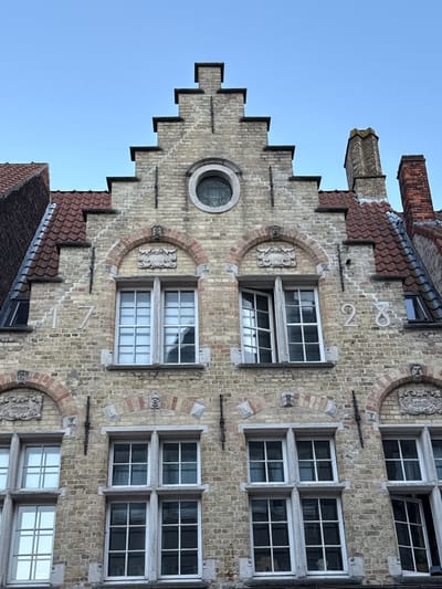 The image shows the facade of a historic brick building in Bruges, Belgium. The architecture is characteristic of Bruges, with stepped gables and ornate brickwork. Several windows of various sizes and styles are visible, some with multiple panes. Two large numbers, "17" and "28", are prominently displayed on the brickwork. A round window with decorative glass is situated near the top of the central gable. Chimneys rise from the roofline. The sky is clear and blue, suggesting good weather. The building appears to be well-maintained, with a sense of age and history.