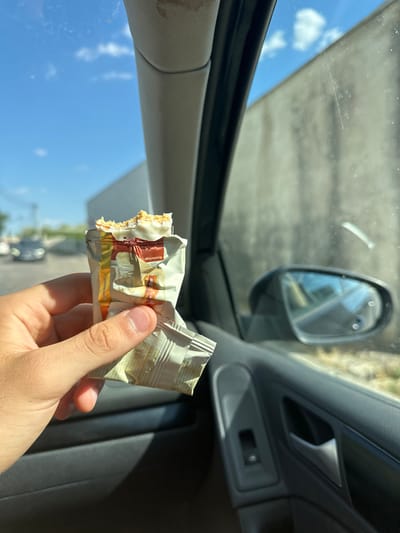 A person's hand holds a partially eaten white chocolate bar inside a car. The hand is positioned in the foreground, obscuring the lower part of the car's interior. The wrapper of the candy bar is silver and white with some red and brown elements. Visible text on the wrapper includes "COLATE BLANC" and "PROTEIN". The background shows the car's interior, including the dashboard, door panel, and side mirror reflecting the sky and trees. The exterior view through the car window reveals a bright blue sky with scattered clouds, suggesting daytime. The scene appears to be in a sunny location, possibly a parking lot or on a road, given the reflections in the side mirror and the blurred foliage outside. There are no discernible emotions conveyed in the image.