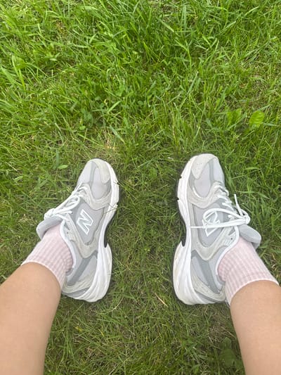 The image shows a pair of grey New Balance sneakers with white laces and a white sole, worn by a person with light skin wearing pale pink socks. The sneakers are positioned on a patch of green grass. The viewpoint is from above, looking down at the person's feet and the surrounding lawn. There are no identifiable landmarks or features to suggest a specific location in Gatineau, Canada. The scene is outdoors, with the grass appearing healthy and well-maintained, typical of a park, backyard, or sports field.