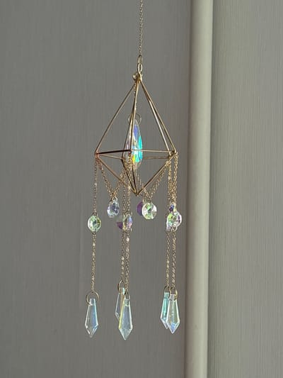 A gold geometric pendant with iridescent crystal accents is suspended against a plain grey backdrop. The pendant features a diamond-shaped frame at the top, from which multiple gold chains descend. Each chain is adorned with small, faceted, iridescent beads and larger, clear, pointed crystals at the ends. The overall lighting suggests an indoor setting, possibly near a window, as indicated by the soft illumination and the presence of a light-colored vertical element on the right, which could be a curtain or blinds. There is no indication of the city of Radkevshchina, Russia, or any people in the image.
