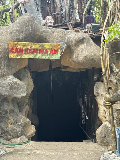 The image shows the entrance to a cave-like attraction, likely a haunted house or similar theme, in Hoa Lư Ward, Vietnam. A weathered sign above the entrance reads "CĂN HẦM MA ÁM" in red lettering against a distressed yellow background, translating to "Haunted Chamber." The entrance itself is a dark, cavernous opening framed by rough, grey rock formations.
Above the entrance, a macabre scene is depicted. A grotesque, disembodied head with wide eyes and an open mouth rests on the rocky overhang. Tattered white fabric, resembling cobwebs or spectral attire, drapes down from above, adding to the eerie atmosphere. A large, rusty chain hangs to the left, further enhancing the spooky theme. To the right, a tombstone-shaped sign with "R.I.P." in red letters is visible, along with a floodlight.
In the foreground, a statue of a warrior figure, elaborately decorated in gold and dark tones, stands to the right of the entrance. The statue holds a long staff. The ground leading to the entrance is paved with textured concrete tiles. Lush green foliage and palm fronds are visible in the background, suggesting an outdoor setting, possibly in a park or tourist area within Hoa Lư Ward. The overall impression is of a deliberately unsettling and theatrical environment designed to evoke fear and amusement.