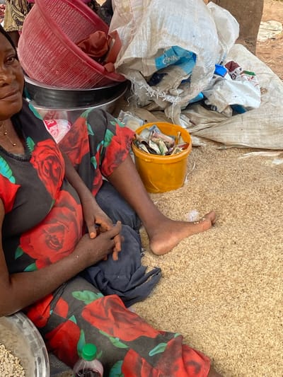 An adult female with a dark complexion, displaying a visible abdominal bulge, is seated on the ground in an outdoor market setting in Igbara, Nigeria. She wears a black garment patterned with large red roses and green leaves. Her hands are clasped in front of her. Her bare left foot rests upon a dense layer of light brown, unhulled rice grains that completely cover the ground surface; several grains adhere to her foot. Her gaze is directed slightly upwards, with a neutral to slightly solemn expression. To her lower left, a partial view of a silver metallic bowl containing light-colored beans is visible. A small clear plastic bottle with a green cap and dark liquid contents is positioned near her right thigh. Behind her, a red perforated plastic basket is stacked on two dark metallic bowls. To her right, a yellow plastic bucket is filled with folded paper currency of various denominations. The background features several large, white woven polypropylene sacks, some appearing partially filled or discarded, with blue text visible on one. A brown earthen structure or pillar is present in the far right background.