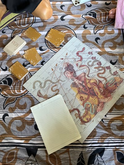 The image shows a close-up, top-down view of a board game and several decks of cards laid out on a patterned fabric. The game board features a central image of a woman entwined with snakes, surrounded by numbered spaces with Russian text. Several stacks of square, gold-colored cards with similar designs are placed to the left of the board, along with one rectangular white box that also contains cards. A blank, lined notepad is positioned in the lower right quadrant of the image, partially covering the game board. The fabric beneath the objects has a repeating pattern of brown and black wavy lines interspersed with thin white lines. In the upper portion of the image, a person's bare leg and knee, along with a black garment, are visible, suggesting they are seated or lying on the fabric. The lighting is consistent with natural daylight. The specific location in Weligama, Sri Lanka, cannot be determined from the visual information present.
