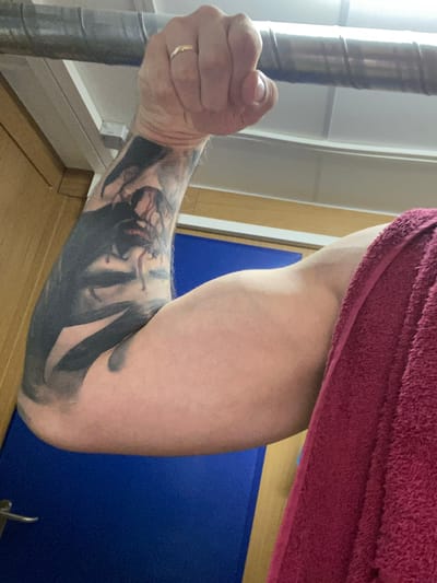 An overhead shot captures a person's left arm, tattooed with a dark, abstract design, gripping a textured metal bar. The arm is flexed, revealing a well-defined bicep, and is partially covered by a plush, maroon towel. In the background, a light-colored ceiling and a light brown wooden cabinet are visible, along with a glimpse of a bright blue surface, possibly a door or wall. The scene is indoors, with fluorescent lighting overhead. The location is not discernible from the image.