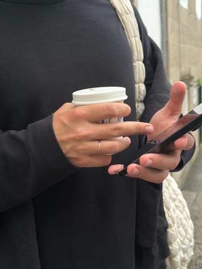 This image shows a close-up view of a person's hands interacting with a smartphone and a disposable coffee cup. The person is wearing a black long-sleeved garment and a gold ring on their left hand. The background appears to be an outdoor setting, possibly a street or a public walkway, with a blurred background that suggests a textured surface, like stone or concrete, and a hint of greenery, possibly indicating a park or garden. The lighting suggests it is daytime, but the absence of strong shadows does not clearly indicate the time of day.
The person is holding a black smartphone in their right hand, seemingly in the process of using it, possibly scrolling or typing. Their left hand is holding a white disposable coffee cup with a white lid, and their fingers are wrapped around it. The cup has some faint blue text visible, though it is largely obscured. The focus is on the hands and the objects they are holding, with the background out of focus. The overall impression is one of casual everyday activity, possibly in an urban environment.
The context provided specifies Santiago de Compostela, Spain. This information, combined with the visual cues of what appears to be an urban setting, suggests the scene could be set in this historic Spanish city. However, there are no explicit landmarks or distinct architectural features visible in the image to confirm the location. The weather appears to be mild, as suggested by the clothing.