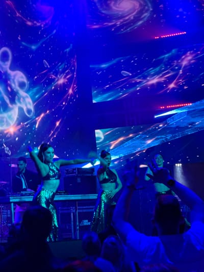 A full shot captures a vibrant concert scene, illuminated by intense blue and purple lighting, with a cosmic-themed projection on the screens behind. In the foreground, the backs of several audience members are visible, with one person in the center raising their arms. On stage, three women in sequined outfits are performing, one in the center with her arms outstretched and the other two on either side in dynamic poses. A man in a black jacket and white shirt is visible on the left behind a DJ booth, with DJ equipment on display. The overall atmosphere suggests an energetic and visually striking musical performance.