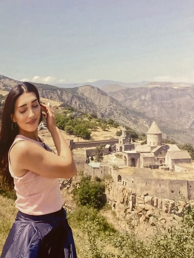 This is a portrait of a young woman posing in front of a mountainous landscape with ancient stone buildings. She is lying on her side, with her head resting on her arm and her eyes closed, a serene expression on her face. She is wearing a light pink tank top and dark blue pants. The background shows a vast, rugged mountain range with a settlement of stone structures built on a hillside. The architecture appears old and possibly historical, with visible courtyards, walls, and a distinct conical tower. Green vegetation is scattered across the slopes. The scene is bathed in bright sunlight, suggesting it is daytime. The clear sky indicates fair weather. The overall impression is one of peacefulness and appreciation for the historical and natural beauty of the location, which is identified as Verin Artashat, Armenia. There is no visible text in the image.