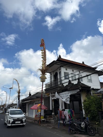 The image captures a street scene in Tampaksiring, Indonesia, under a partly cloudy blue sky. A white car, license plate DK 1451 PO, is driving down the asphalt road on the left. To the right of the car, a tall, ornate Balinese pole, likely a Penjor, stands adorned with dried leaves and decorative elements. Further down the street, other similar poles with curved tops are visible.
Buildings line the right side of the street. One building, a white two-story structure with a dark tiled roof, features a balcony and several windows. In front of another single-story building, clothes are hung on a clothesline supported by colorful poles. A black motorcycle is parked on the sidewalk. A red umbrella is set up, and a table displays various items. A banner hangs on the front of the building, featuring text in Indonesian. Electrical wires are strung across the scene, crisscrossing overhead. Lush green foliage is visible on the right side of the image.