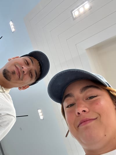 Two individuals are captured in a close-up selfie within an interior space. The ceiling above is visible, featuring a white herringbone pattern with recessed rectangular lighting fixtures. The person at the top of the frame is wearing a blue baseball cap and has light skin, while the individual at the bottom also sports a blue baseball cap and has a goatee. The location may be in the city of West Hollywood, United States of America. No specific actions or interactions are discernible beyond the act of taking a selfie. The scene is dominated by the faces of the two people and the overhead ceiling.