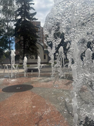 This is an upside-down, close-up photograph of a fountain in Stupino, Russia. The main subjects are the water spraying from the fountain and the textured, reddish-brown surface of the fountain's base. The water is in motion, with many droplets and bubbles visible, catching the light and creating a dynamic texture. In the background, blurred reflections in the water show trees and a multi-story building, suggesting an outdoor setting with some urban elements. A dark, circular grate is visible on the fountain's base, with a white ring in its center. The lighting suggests it is daytime, possibly sunny, given the bright reflections on the water. The image's orientation and the close proximity to the water create a sense of immersion and perhaps a feeling of refreshment. No people are visible in the frame.