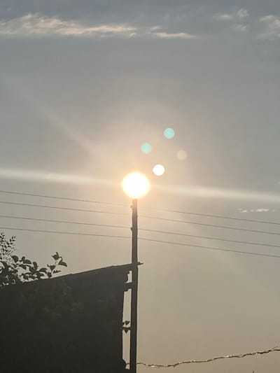 The image depicts a streetlamp against a bright sky, with the sun visible and causing lens flare. The streetlamp is a dark pole with a round, illuminated light fixture at the top. There are power lines strung horizontally across the frame. In the foreground, a dark, silhouetted structure, possibly a building or fence, is visible on the left, with some dark foliage in front of it. Several circular orbs of light, likely reflections or lens flares, are scattered in the sky around the sun. The sky itself is a hazy gray with some wispy clouds. The scene appears to be an outdoor, possibly urban or semi-urban, setting. The image was taken in Rakity, Russia.
