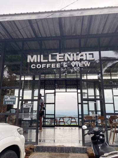 The image shows the entrance of a coffee shop called "Milleniad Coffee & View" located at the coordinates -7.855, 110.473 in Kecamatan Piyungan, Indonesia. The building is made of black metal framing with large glass windows. An "OPEN" sign is hung on the door. A man wearing a white shirt and black shorts is standing in the doorway. He is facing the viewer. A black scooter is parked in front of the doorway. The scooter's handlebar is visible. The interior of the coffee shop is visible through the glass doors. There is a small table and chairs placed in front of the door on the left side of the image. The exterior of the shop is on the right side of the image. It is a large, open area with a view of distant mountains. The sky is cloudy and overcast. There is a sign that says "BALKON BAWAH LUAS" which translates to "lower balcony spacious" hanging on the left side of the door frame. The sign is written in Indonesian. The exterior wall of the building is made of brick.