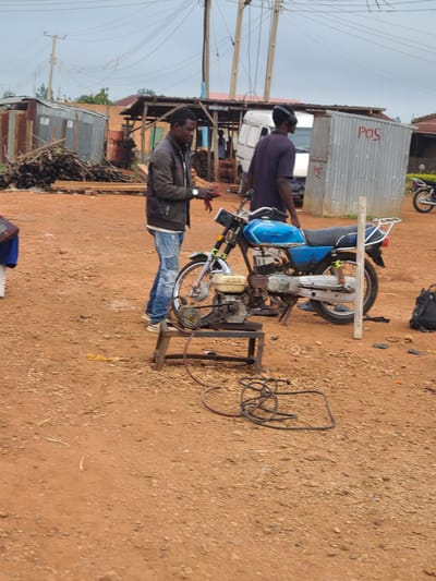 The image depicts an outdoor scene in Barikin Sabon Gidan Kanat, Nigeria. In the foreground, a blue and silver motorcycle is parked on reddish-brown dirt. A portable generator is situated in front of the motorcycle, connected by wires to something out of frame. Two men are present. One man, wearing a dark jacket over a white shirt and blue jeans, stands to the left of the motorcycle, appearing to be working with something red in his hands. The other man, wearing a dark purple shirt and black headphones, is partially visible behind the motorcycle, facing away from the camera. In the background, various structures are visible, including a corrugated metal shed with "POS" written in red letters, a wooden stall, piles of logs, and a white van. Electrical poles and overhead wires are prominent in the sky. The overall environment suggests a semi-urban or market area, possibly a workshop or service point.