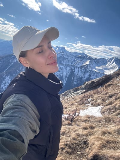 A person wearing a light gray baseball cap, a gray long-sleeved shirt, and a black puffer vest is positioned on a mountain ridge, likely in a selfie perspective. The person's eyes are closed, and they are facing towards a partly cloudy sky with bright sunlight. Behind the person, there is a steep, rocky, mountainous terrain with patches of dry brown grass and snow. In the distance, snow-capped mountains are visible under a clear blue sky. The overall scene suggests a location high in the mountains, with signs of late autumn or early spring weather. The photo was taken in Arshan, Russia.
