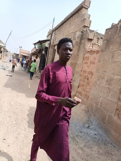 A young adult male, of dark complexion with short black hair, stands in the foreground, angled slightly to his right (viewer's left), looking directly forward with a neutral expression. He wears a long-sleeved maroon traditional garment consisting of a tunic with a mandarin collar and matching trousers. His left hand holds a small, light brown, rectangular object. He is positioned on a dry, unpaved dirt path. To his left (viewer's right) is a damaged light-colored concrete block wall, showing a jagged top edge and exposed internal structures. In the midground, further down the dirt path, several individuals are visible: one person in a light brown garment walking away, another in a bright green shirt, and additional figures in the distance. Bicycles are present among the people. Simple buildings with various roofing materials, including corrugated metal, line the left side of the path. Overhead utility lines are visible. The sky is bright and clear. The scene depicts a street in Zango, Nigeria.