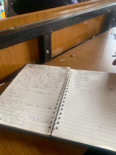 This media file captures a close-up view of an open notebook lying on a wooden surface, likely a desk or table. The notebook is spiral-bound and contains handwritten notes in English, with some discernible text including "Nigerian Home Videos," "1933," and "Evolution of Nigerian Home Videos 1903-1984." The handwriting suggests a personal note-taking session, possibly for academic or research purposes. The notebook is angled, with the left page mostly visible and the right page partially obscured. The wooden surface appears to be a light-colored wood, possibly a desk, with a dark metal frame visible along the left edge of the frame. The lighting suggests it is daytime, and there are no clear indications of weather conditions or specific location cues beyond the textual content within the notebook which mentions "Nigeria." The overall impression is one of focused study or research.
