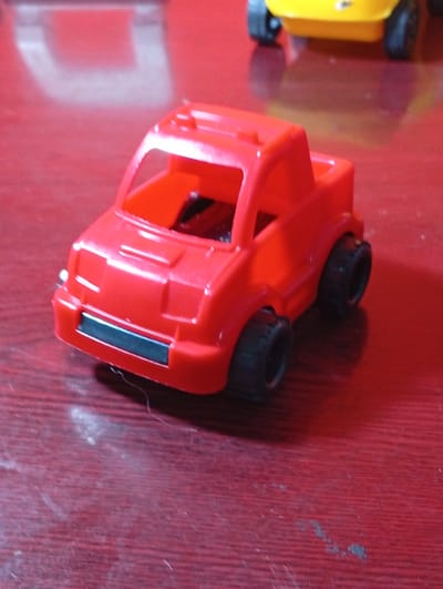 A red plastic toy truck is positioned on a red, wooden surface. The truck is facing the left, with its front bumper slightly angled. It has black wheels. In the background, out of focus, is a yellow toy vehicle. The surface appears to be a table. This scene is not located in Khujayli, Uzbekistan; it is a general object photograph.