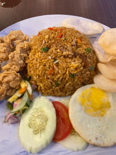 This image shows a plate of food, likely a meal. The main subject is a mound of fried rice, which appears to be mixed with various ingredients such as small pieces of meat, vegetables like carrots and green onions, and possibly egg. To the left of the rice, there are several pieces of what look like crispy fried chicken. To the right of the rice, there is a fried egg with a runny yolk, a slice of tomato, and a slice of cucumber. A small portion of a fresh vegetable salad, possibly a pickle or acar, with thinly sliced cucumber, carrots, and red onion, is visible at the bottom. Several crispy crackers, likely krupuk, are placed above the fried rice. The food is served on a white plate with a blue, marbled pattern. The setting appears to be a table, with a dark wooden surface visible in the background, suggesting an indoor dining environment. The lighting suggests it could be daytime, as there are reflections on the food and plate, indicating overhead lighting or natural light. There is no visible text in the image. The meal appears to be a savory dish, typical of Indonesian cuisine, and the presentation suggests it is ready to be eaten.