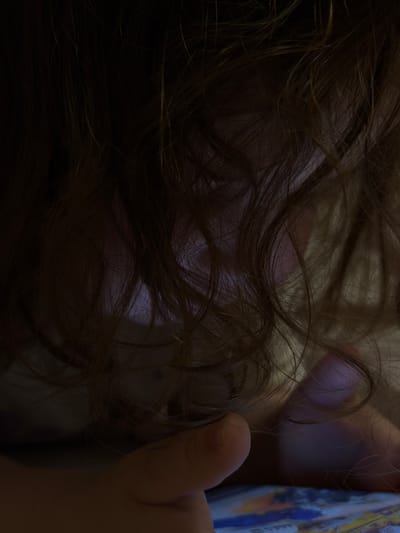 In Bobrovo, Russia, this close-up, low-light image focuses on the tangled, dark brown hair of a child. The child's head is bent down, obscuring their face, but their hands are visible in the foreground, one of which is resting on a colorful, patterned surface that appears to be a tablet or a book. The lighting suggests the child is engaged with a screen, which is casting a faint purple glow through their hair. The background is dark and indistinct, emphasizing the texture and movement of the hair.