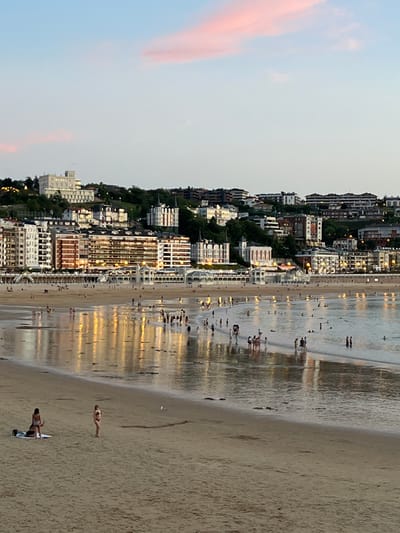 This is a photograph of La Concha beach in San Sebastián, Spain, during twilight. The sky is a pale blue with streaks of pink from the setting sun. The city skyline is visible in the background, with a prominent white building atop a hill, likely a hotel or historical site, followed by rows of multi-story buildings with illuminated windows. The beach is populated by numerous people, some walking along the shore, others wading in the shallow water. The wet sand reflects the lights from the city buildings, creating a shimmering effect. In the foreground, two women are visible on the sand, one sitting and the other standing, both in swimwear. The overall scene depicts a lively evening at a popular urban beach.