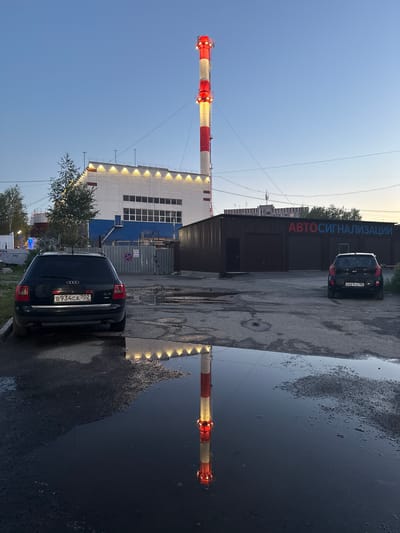 In Surgut, Russia, a reflection of a tall, red and white striped industrial chimney with red lights at the top is visible in a large puddle on a dark, asphalt surface. The chimney rises against a clear, twilight sky. In the background, a modern white and blue industrial building with illuminated windows runs horizontally. To the right, a dark-colored commercial building with two large garage doors and the red Cyrillic text "АВТОСИГНАЛИЗАЦИИ" (AUTO ALARM) is situated. A black Audi station wagon is parked on the left, and a black Kia hatchback is parked in front of the commercial building on the right. The ground is uneven and appears to be a parking lot or service area with scattered debris and smaller puddles.