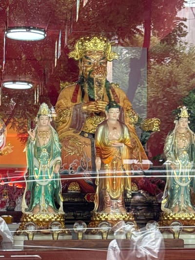 A medium shot captures a display of religious statues behind a reflective glass pane. The central figure is a large, imposing statue of a deity with a golden headdress, beard, and ornate orange and gold robes, its arms resting on its lap. Flanking this central figure are two smaller statues of what appear to be deities or bodhisattvas, one on the left and one on the right. The statue on the left wears a green and gold robe and a tall, ornate headdress. The statue on the right also wears a green and gold robe and a headdress. In front of these statues, another smaller statue of a figure in golden and orange robes with a swastika symbol on its chest stands between the larger central figure and the right-hand flanking figure. The statues are displayed on golden pedestals, which rest on a multi-tiered wooden platform. The background is a deep red, possibly a wall or fabric, with decorative elements and illuminated panels hanging from the ceiling. Reflections in the glass obscure some of the background details, showing trees and buildings outside. The image is likely from Linghe, China, given the context provided.