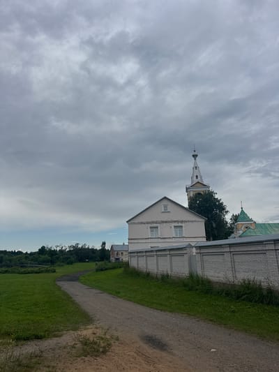 The image captures a view of a pathway leading towards a walled compound in the city of Рассвет, Russia. The sky is overcast with dense, gray clouds. To the right, a white brick wall borders the property, behind which stands a pale pink building with white-trimmed windows. Beyond this building, a prominent bell tower with a white spire and yellow accents rises into the sky, partially obscured by lush green trees. To the left of the pathway, a vast expanse of green grass stretches towards a line of dense trees and foliage. In the distance, a pedestrian can be seen walking along the pathway, and a rustic wooden bridge is visible crossing a body of water within the greenery.