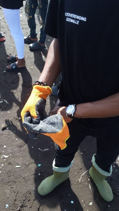 The image shows a close-up of a person's hands and feet, holding a pair of orange and black gloves. The person is wearing black pants, green rubber boots, and a black t-shirt with white writing on it. The writing says "LSVERENIGING EDEWALE". The person is standing on a dirt road in Limbe, Cameroon. The coordinates 4.004, 9.211 place the location near a beach area. The person's hands are visible, the right hand holding the gloves and the left hand is obscured from view. The image appears to be taken from a first-person perspective. The subject is standing, possibly looking at the gloves, the only visible part of their body. The background is a blurred image of people walking on the same dirt road. The image is focused on the gloves and the person's hands. The environment is a dirty, outdoor area with a dirt ground and some scattered debris. The image depicts the act of putting on gloves, which is a common action when engaging in activities such as gardening or construction, but the details of the setting and the location suggest a more informal context. The specific location of the photo, near the beach, and the presence of other individuals suggests a possible connection with coastal or beach cleanup activities. However, this can only be a speculation, and the context of the image remains somewhat ambiguous.