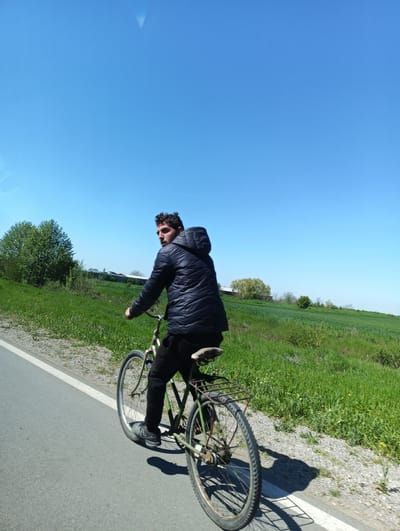 A person is riding a bicycle on a paved road. The person is wearing a dark jacket and dark pants. The bicycle appears to be old and has a rack on the back. The road is bordered by a strip of grass and gravel on one side, and a field of green grass and vegetation on the other. The sky is clear and blue. This photograph was most likely taken outside of the city limits of Straldzha, Bulgaria, as the environment shows a mostly natural landscape.