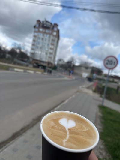The image shows a cup of coffee with latte art in the foreground, held by a person. The background is a blurred street scene. A building is visible on the left, and a road sign with the number "20" is on the right. The setting appears to be Sîngera, Moldova, based on the street architecture. The weather seems cloudy.