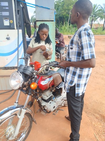 The scene depicts a fuel station in Adikpo, Nigeria, set on reddish-brown dirt ground with green foliage and distant structures under an overcast sky. In the foreground, a male subject wearing a short-sleeved blue, black, and white plaid shirt and dark trousers stands beside a red and chrome motorcycle, holding its handlebars. To the immediate left of the motorcycle, a female subject with braided dark hair, wearing a light-colored short-sleeved top, is intently counting a stack of banknotes held in her hands. A white and blue fuel dispensing pump is partially visible to the far left. A second male subject is partially visible in the mid-ground background, standing near another motorcycle to the right of the fuel pump.